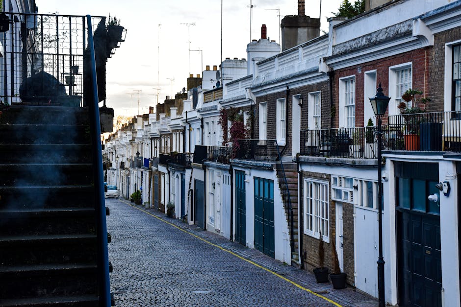 A quiet residential street in Walthamstow with a row of terraced houses featuring brick and painted facades, small front gardens with potted plants and decorative elements, and traditional black metal balconies along the upper levels. The scene shows a cobblestone pavement with a parked white van at the end of the street, situated near a staircase with dark steps and black metal railing on the left. The houses have white-framed windows and chimneys extending from the rooftops, with some windows displaying curtains or blinds. The street is illuminated by natural light likely from late afternoon or early evening, and a lamppost is visible on the right side. This setting provides a typical backdrop for home relocation or furniture transport activities, where [COMPANY_NAME] might be assisting with packing and loading in the context of house removals in Walthamstow, as described in the Hoe Street moving guide.