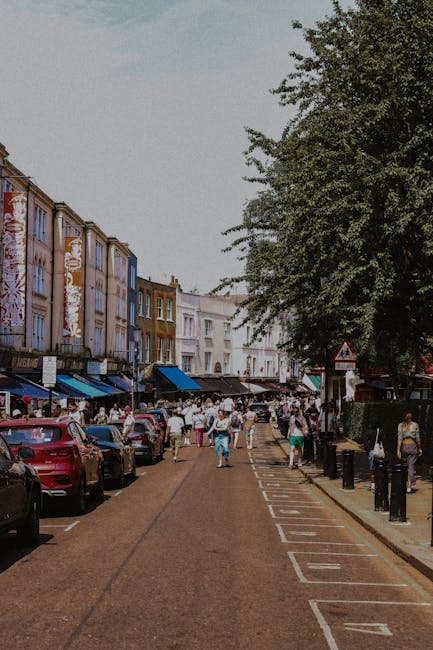 A quiet residential street in Walthamstow with a row of terraced houses featuring brick and painted facades, small front gardens with potted plants and decorative elements, and traditional black metal balconies along the upper levels. The scene shows a cobblestone pavement with a parked white van at the end of the street, situated near a staircase with dark steps and black metal railing on the left. The houses have white-framed windows and chimneys extending from the rooftops, with some windows displaying curtains or blinds. The street is illuminated by natural light likely from late afternoon or early evening, and a lamppost is visible on the right side. This setting provides a typical backdrop for home relocation or furniture transport activities, where [COMPANY_NAME] might be assisting with packing and loading in the context of house removals in Walthamstow, as described in the Hoe Street moving guide.