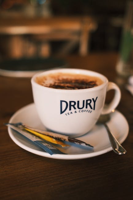 A close-up view of a white ceramic cup filled with hot coffee, topped with a layer of frothy foam and a sprinkle of cocoa powder. The cup, branded with the 'DRURY TEA & COFFEE' logo, rests on a matchi